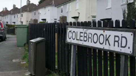 A street sign with the words 'Colebrook Road' in front of a black fence around a garden. There is a pavement in front of the fence with a green wheelie bin and houses on the right. 