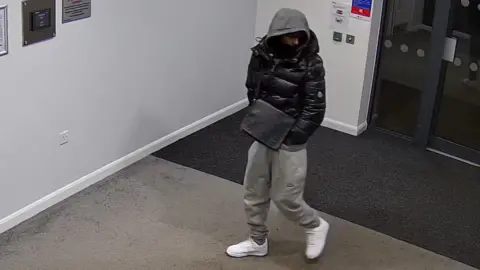 Dualla Abdi walks through the entrance of a building wearing grey tracksuit bottoms, white trainers, a black jacket and his grey hood pulled over his head. The Louis Vuitton bag is black and is worn across his body as a shoulder bag. 