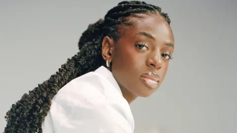 Reuben Bastienne Cat Burns in a promotional shoot. She wears a white shirt and has her braided hair tied back. She looks at the camera with a serious expression over her right shoulder. She's pictured against a grey backdrop. 