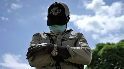 James (not his real name) looks down as he poses for a photo with his arms folded wearing a wrist watch, baseball cap, face mask, trousers and a khaki shirt. He is standing outside and a blue sky can be seen in the background