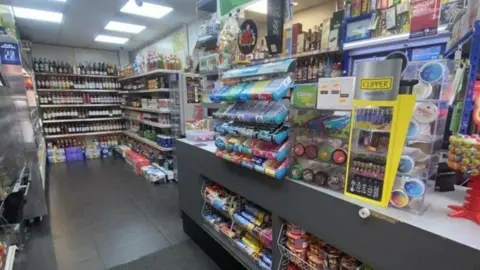 Ernest Wilson Business Agents The shop counter is filled with products for sale including chewing gum and biscuits. Shelves further back are filled with products including bottles of wine.