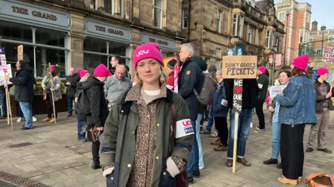 BBC Jo Grady stands in front of a group of picketers. She is wearing a pink hat that reads: "UCU". Behind her, a placard reads: "Don't cross pickets".