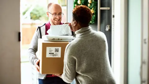 Getty Images A bald delivery man, wearing a red t shirt and grey zip-up top hands over cardboard box to female customer, at her door. She is only seen from the back, and is wearing a grey jumper and has short, curly dark hair. There is a Christmas wreath on the door.