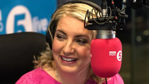 Anna Foster pictured in front of a microphone in the BBC Radio 5 Live studio. She is wearing a pink top.