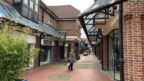 Phil Harrison/BBC An outdoor street in a shopping centre. There is a woman in a grey coat walking in the red bricked-alley.