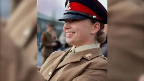 Family Handout Jaysley Beck wearing her brown formal military uniform and a black and red cap. She has her light brown hair tied back into a low bun and is looking off to the left of the camera and smiling.