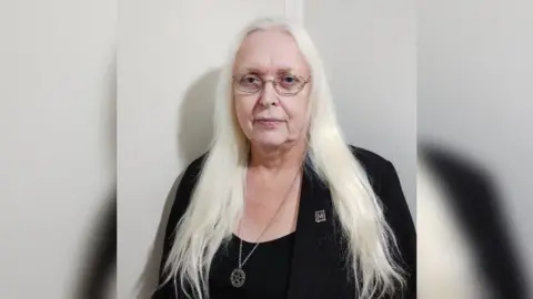 A formal mid-shot of Maz, who has long white hair and glasses, and is wearing a black vest and jacket