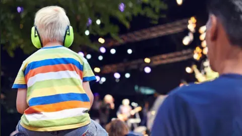 Generic shot of the back of a young boy wearing a stripy t-shirt sitting on the shoulders of an adult. They are facing a stage where a band is playing - this is in the background and is out of focus.