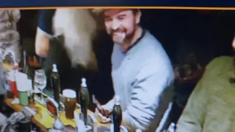 Dyfed-Powys Police  a man with a beard in grey jumper smiling broadly as a he sits at the bar with the beer taps in front of him. He is also holding a mobile phone