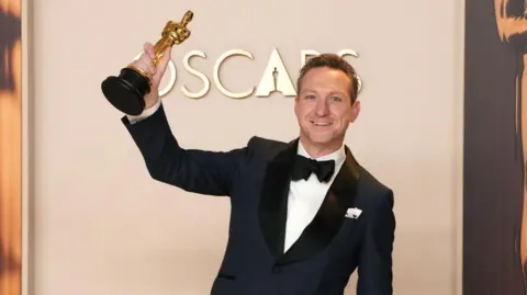 Lol Crawley, winner of the Best Cinematography for “The Brutalist”, poses in the press room during the 97th Annual Oscars at Dolby Theatre on March 02, 2025 in Hollywood, California. He is wearing a navy blue suit with a black bow tie and a white shirt. He is holding the Oscar in his right hand in the air. Behind him is a sign that says "OSCARS"