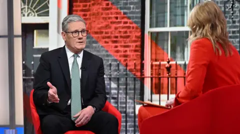 Sir Keir Starmer sits on a red chair opposite Laura Kuenssberg and is talking. He is wearing a white shirt, mint-green tie, and black blazer. He has grey hair and is wearing black-rimmed glasses. Laura Kuenssberg is wearing a red blazer and trousers, and she has blonde hair.