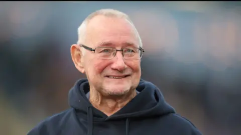 A man with white hair and glasses, wearing a blue hoody