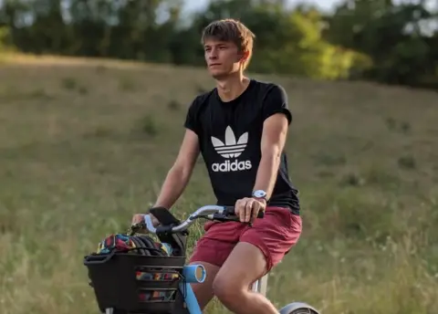 Nathalie Tison A young man wearing an adidas top cycles on a blue bike, with a field in the background