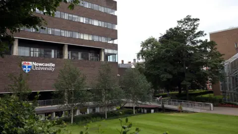 Newcastle University's Claremont Tower. The dark brick building is several storeys high and has the university logo on one wall. It is surrounded by grass and trees.