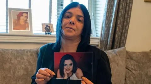 A black haired woman in a black top is holding a photo of her daughter towards the camera. The girl in the photo is smiling. The woman is sitting on a beige sofa with a window behind her.