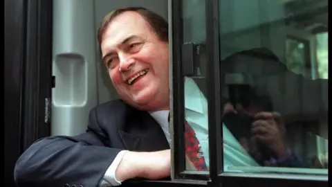  John Prescott wearing a black suit and hanging out of a bus window 