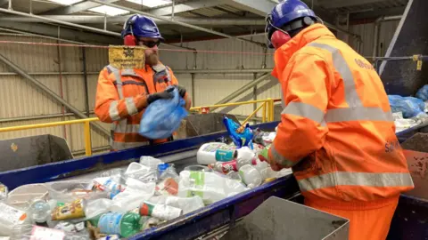 Two men in high vis uniform standing by a conveyor belt sorting mixed plastics