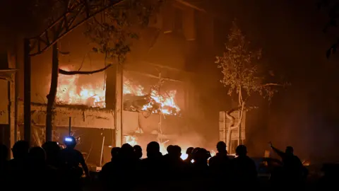 EPA A building is in flames at night, with a crowd of firefighters and onlookers below