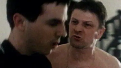 Granada Films A topless Sean Bean is talking angrily at another man, who has black hair and, although slightly blurred, looks concerned.