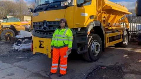 Heidi Tomlinson/BBC A woman who is a gritter driver dressed in orange and yellow hi-vis stands next to a gritter 