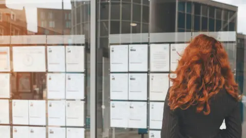 Getty Images A woman with long red hair stands in front of a bulletin board with job listings, reviewing the possibilities.