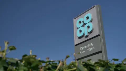 A co-op logo on a sign against a blue sky with a green hedge in the foreground