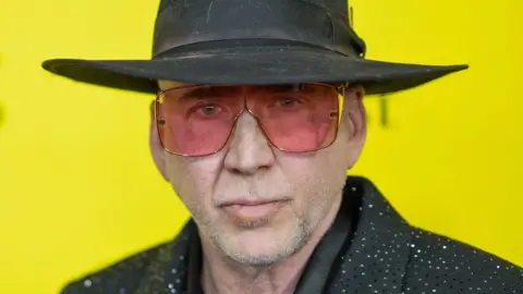 Nicolas Cage looking at the camera in a sparkly black suit, black hat and pink sunglasses, against a bright yellow background at the premiere of The Surfer in Austin, Texas, on 10 March