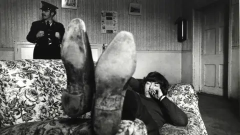 Siobhan O'Sullivan Harvey Brendan is pictured laying on a floral couch as a young man. The bottom of his scuffed up formal shoes are close to the camera on the armrest. Brendan lays back in dark jeans and a jumper with one arm behind his head. He is smoking a cigarette with the other hand, with a watch poking out of his sleeve. He has medium length dark hair which is swooped to the side of his face. In the background of the image a scuffed up white door is to Brendan's right, while a police officer stands to his left. He is behind the couch but in front of a wood panel wall. The officer is dressed in an old style black police uniform. It has large metal buttons and a belt buckle on show. 