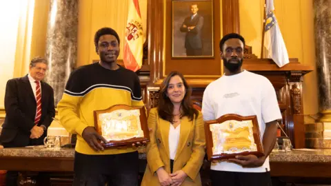 (L-r) Magatte N'Diaye, Inés Rey and Ibrahima Diack in the council hall room. The men holding their plaques stand on either side of the mayor.