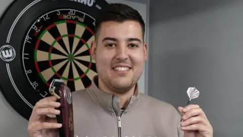 Ryan Meikle smiles at the camera while standing in front of a dartboard. He is holding an electric hair clipper in one hair and a dart in his other. He has short dark hair and is wearing a grey coloured jumper.