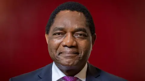 AFP Zambian President Hakainde Hichilema smiles at the camera, wearing a suit and a patterned purple tie.