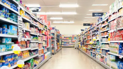 Getty Images Shelves in a grocery store aisle.
The products are blurred out but colourful and the titles at the top of the aisle are blue and "fish and meat" sign is visible. 
The floor is cream and the lights are bright. 