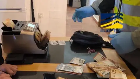 Europol/Spanish Police The hands of two investigators examining a large pile of euro notes on a table. There is also a money counter. The image is watermarked with the logos of the Spanish National Police and Interpol. 