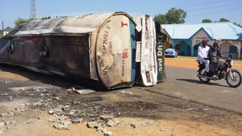 AFP Two people on a motorbike drive by the burnt-out tanker on the side of the road in Majia, Nigeria - 16 October 2024