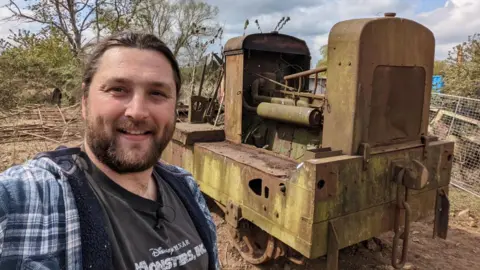 Lawrie takes a selfie style photo and smiles at the camera. He has dark long hair that is tied back as well as a dark beard. He stands in front of an old locomotive in a scrapyard that is rusty and worn.