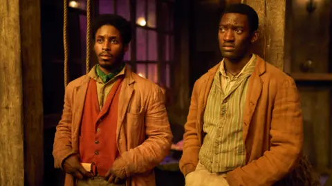 Disney Francis Lovehall (left) Malachi Kirby (right) in costume as their characters Alec Munroe and Hezekiah Moscow. Both are wearing light brown jackets and colourful patterned waistcoats