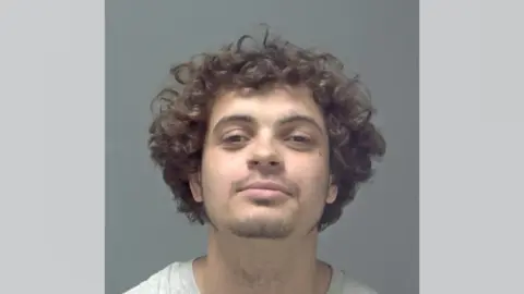 Suffolk Police A mugshot of Logan Burnett, who is smiling at the camera with his head tilted upwards. He has dark, curly hair and brown eyes. He has a small amount of facial hair.
