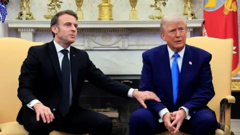 Both seated in the Oval Office, Macron reaches over to place his arm on Trump's arm as he begins speaking