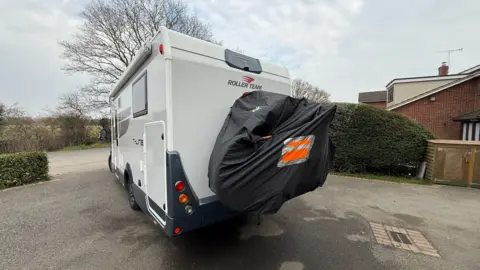 Steve Hubbard/BBC Long shot showing the couple's white modern motorhome with a bike rack attached to the back and concealed with a black zipped cover