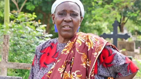 BBC Pudensiana Chumbi, wearing a headwrap and dressed in a flower print dress with long scarf draped over one shoulder, a stands in the graveyard in Morogoro with a hand on her hip looking at the camera.