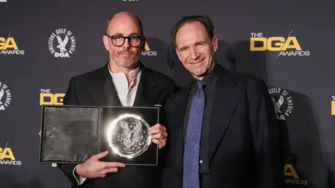 Getty Images Ralph Fiennes and Edward Berger pose for a camera on a red carpet. On the right, Fiennes is wearing a dark suit and blue tie. On the left Berger is wearing a dark suit and white open neck shirt. 