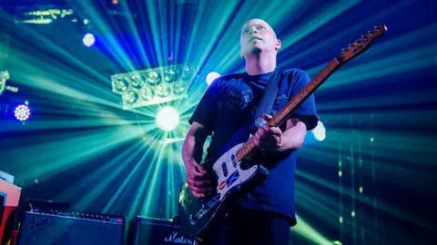 Getty Images Stuart Braithwaite of Mogwai strums his guitar as a spotlight illuminates him from behind