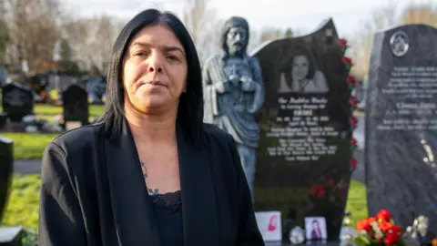 Helen stands in front of her daughter's grave. She is dressed in black and looks at the camera. 