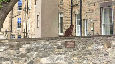 Alan Beattie Herriot A drawing showing how the brown cat would be sitting on the wall. There is a plaque underneath but you can't make out the writing on it. There are tenement flats in the background.