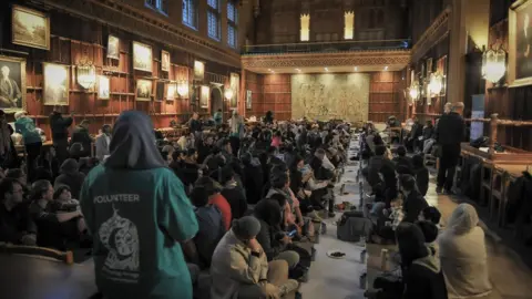 People sitting cross-legged on the floor of the dining hall inside the Cambridge College -breaking their fast 