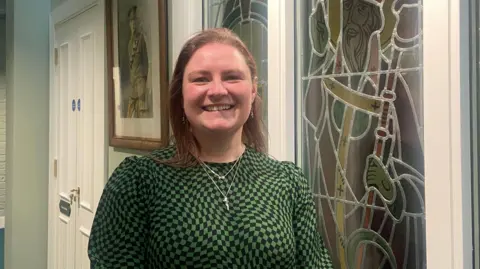 Dr Laura Patrick. She has shoulder-length brown hair, wearing silver necklaces and a green and black dress. She is standing in front of a stain glass window and a military portrait and be seen to her left.