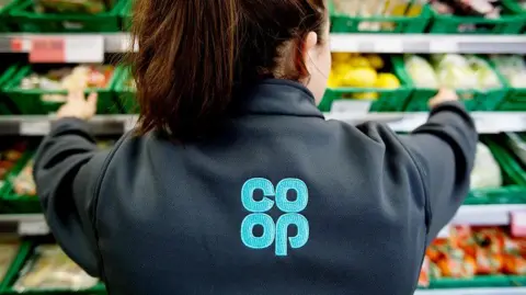 Co-op The back of woman with brown hair in a ponytail wearing a Co-op uniform with the logo on her back while reaching across shelves of vegetables