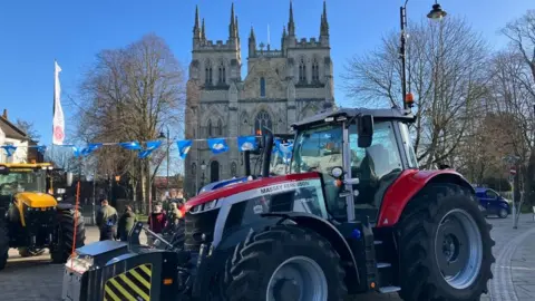 BBC/Mark Ansell Two tractors parked outside a church in a town. In the background there are people and a string of blue flags hanging in the air. 