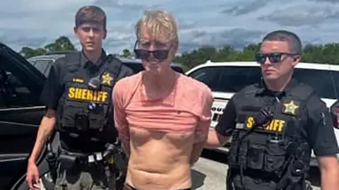 Reuters / Martin County Sheriff's Office A still from a police video showing Ryan Routh, a man with a mop of blond hair, sunglasses and a pink T-shirt pulled up over his stomach, flanked by two deputies from the Martin County Sheriff's Office