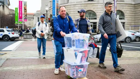 Rick Bielke, a former USAID employee, who believes he is currently on administrative leave but is unsure of his status as he is locked out of his communications, pushes personal items on a skateboard as he leaves the former USAID offices at the Ronald Reagan Building and International Trade Center on February 27, 2025 in Washington, DC.